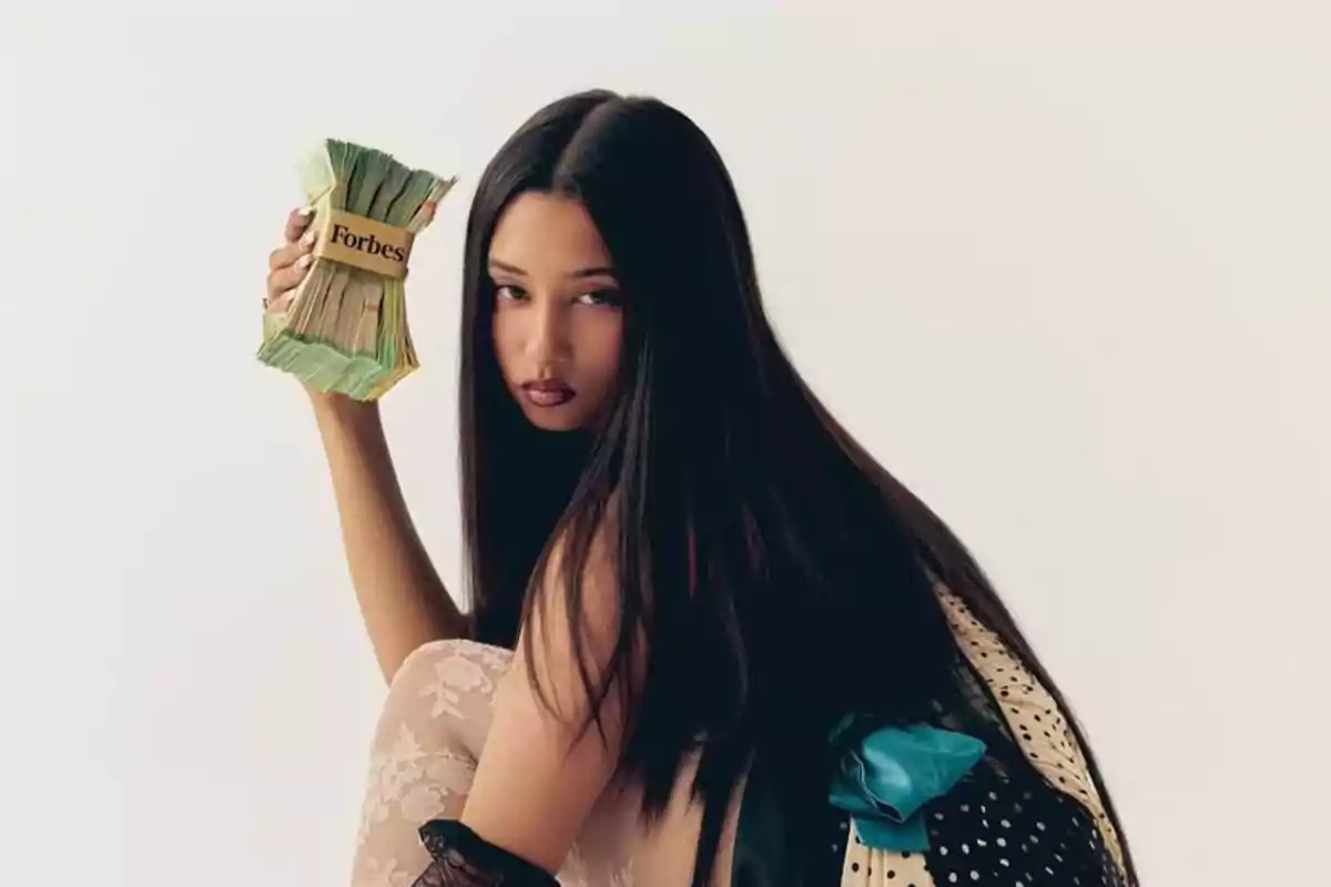 Mujer de cabello largo y lacio sostiene un fajo de billetes con una banda que dice Forbes mientras mira a la cámara