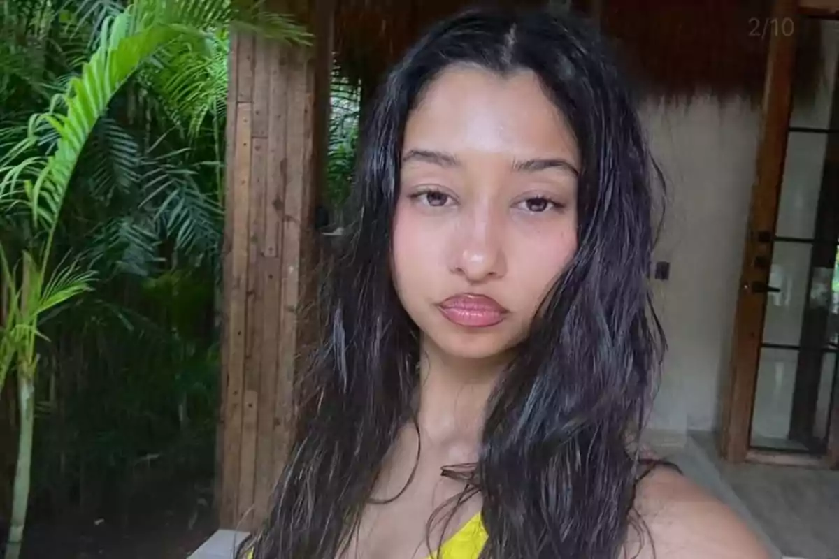 Mujer de cabello largo y oscuro con expresión seria tomando una selfie en un entorno natural con plantas y una estructura de madera al fondo