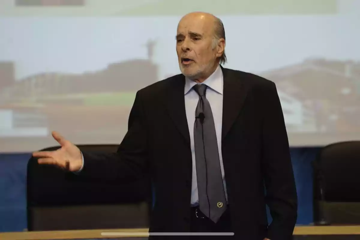 Hombre mayor de traje oscuro hablando frente a una audiencia con fondo de proyección borroso