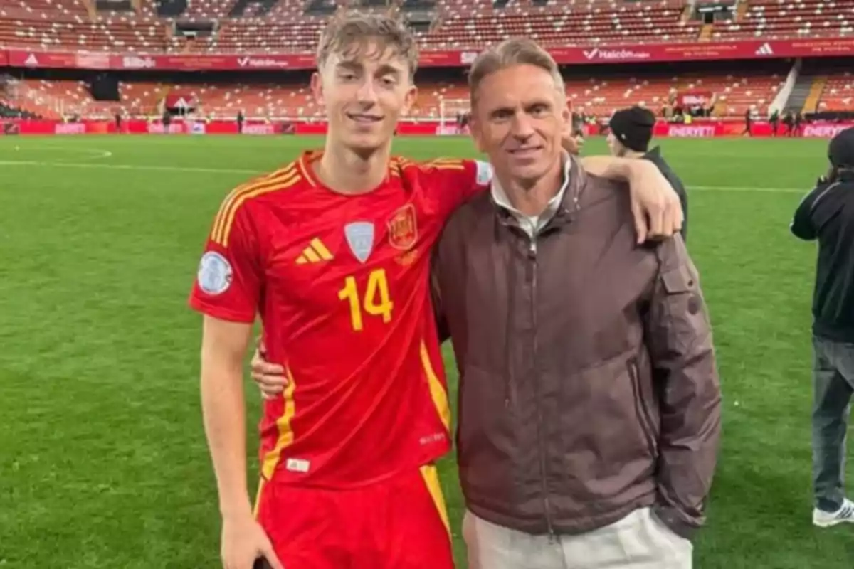 Un futbolista con uniforme rojo y el número 14 posa sonriente junto a un hombre en una cancha de estadio con gradas al fondo