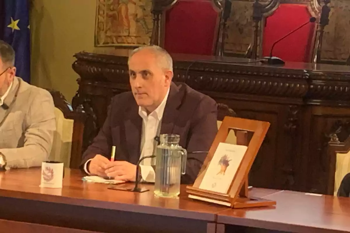 Un hombre de cabello canoso y saco oscuro está sentado en una mesa de madera, sosteniendo un bolígrafo, con una jarra de agua, una taza y un libro en un atril frente a él, en un entorno formal con sillas tapizadas y una bandera de la Unión Europea al fondo.