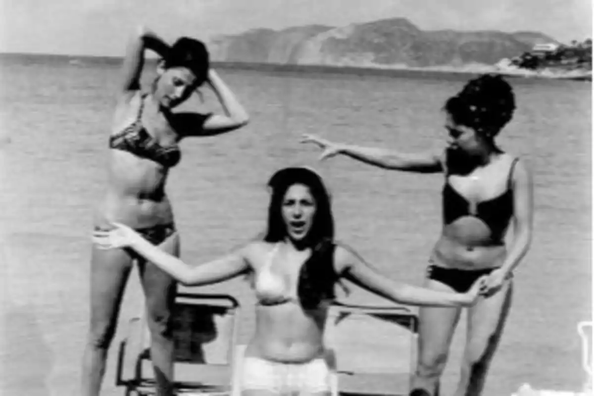 Tres mujeres en bikini posan en la playa con montañas al fondo en una fotografía en blanco y negro Tres mujeres en bikini posan en la playa con montañas al fondo en una fotografía en blanco y negro