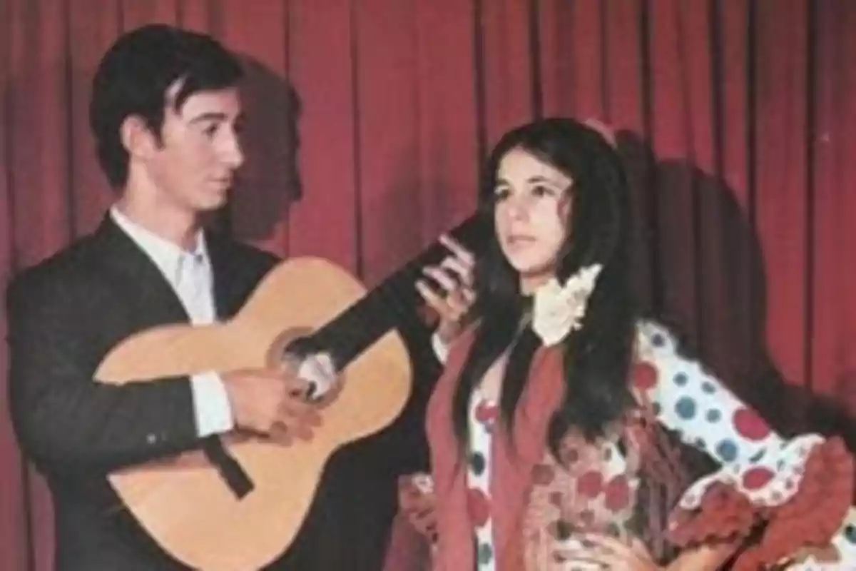Un hombre con guitarra y una mujer vestida con traje de flamenca posan frente a un telón rojo Un hombre con guitarra y una mujer vestida con traje de flamenca posan frente a un telón rojo