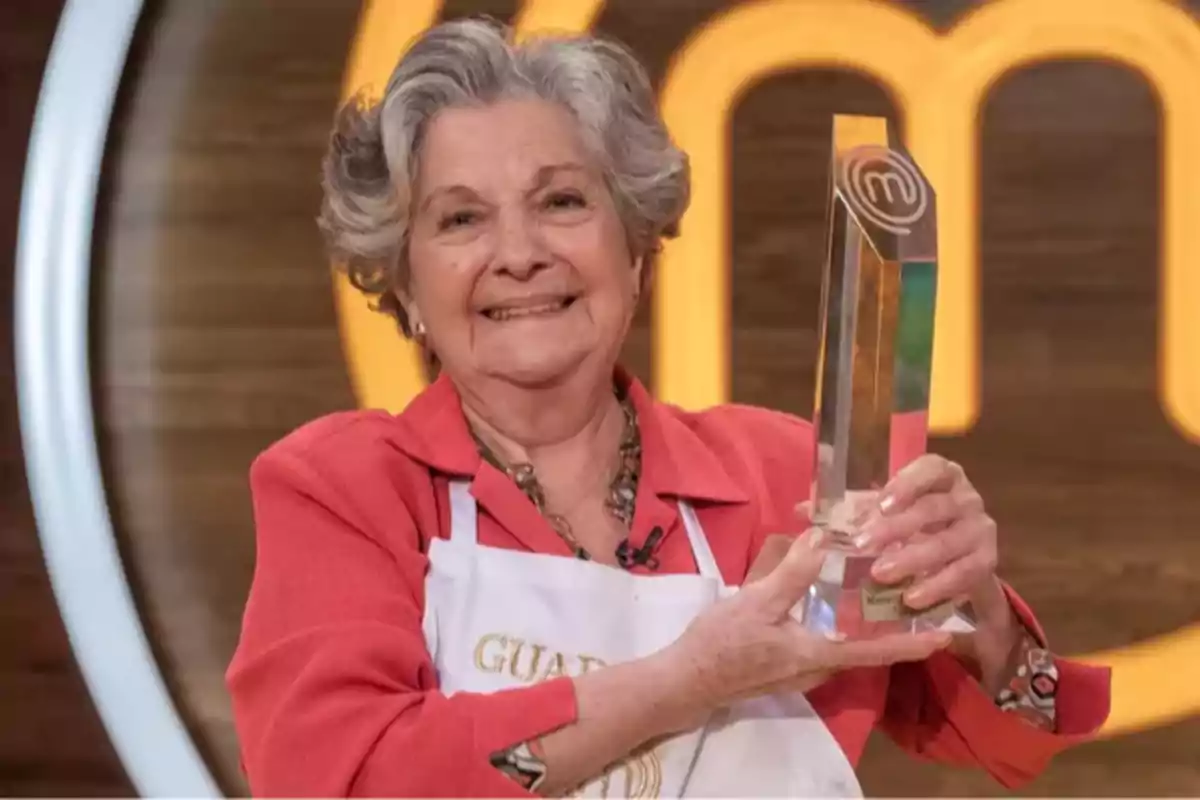 Mujer mayor sonriente con cabello canoso y delantal blanco sosteniendo un trofeo de MasterChef frente a un fondo con el logo del programa Mujer mayor sonriente con cabello canoso y delantal blanco sosteniendo un trofeo de MasterChef frente a un fondo con el logo del programa
