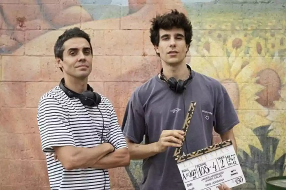 Dos hombres jóvenes con auriculares alrededor del cuello posan frente a un mural colorido, uno de ellos sostiene una claqueta de cine
