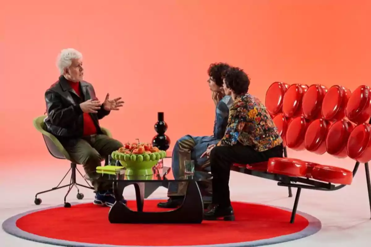 Tres personas conversan en un set moderno con fondo naranja, sentados en sillones de diseño y una mesa con frutas al centro Tres personas conversan en un set moderno con fondo naranja, sentados en sillones de diseño y una mesa con frutas al centro