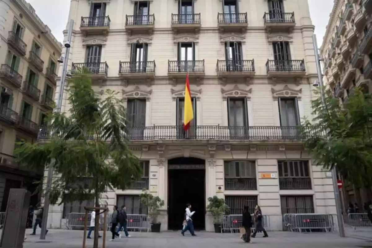 Fachada de un edificio oficial con una bandera de España en el centro y varias personas caminando por la acera