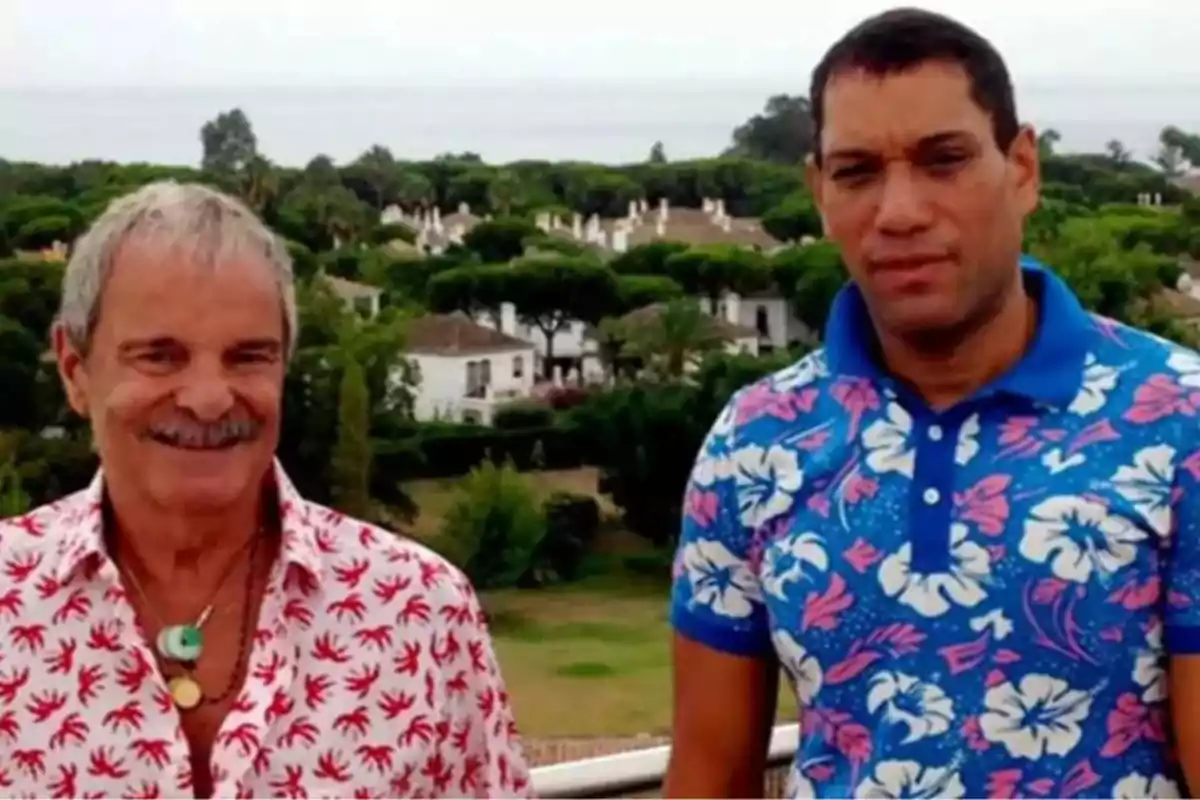 Dos hombres posan juntos al aire libre con un paisaje de casas y árboles al fondo, ambos llevan camisas coloridas con estampados llamativos. Dos hombres posan juntos al aire libre con un paisaje de casas y árboles al fondo, ambos llevan camisas coloridas con estampados llamativos.