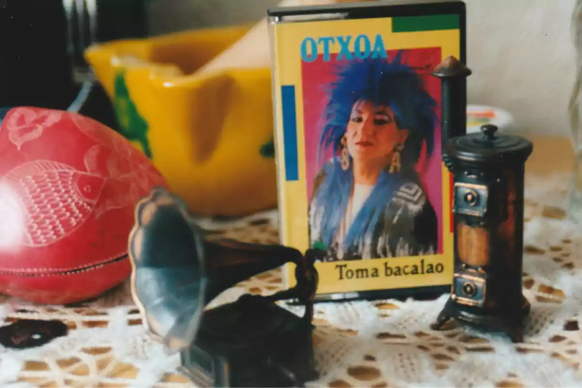 Cassette de Otxoa titulado Toma bacalao sobre una mesa con objetos decorativos como un gramófono en miniatura, una figura de estufa y una pieza roja con grabado de pez Cassette de Otxoa titulado Toma bacalao sobre una mesa con objetos decorativos como un gramófono en miniatura, una figura de estufa y una pieza roja con grabado de pez