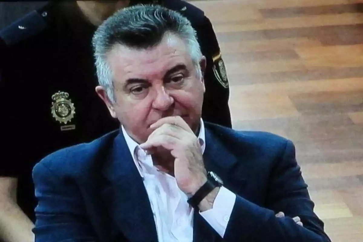 Hombre de cabello canoso con expresión pensativa sentado en una sala con piso de madera y un policía al fondo Hombre de cabello canoso con expresión pensativa sentado en una sala con piso de madera y un policía al fondo