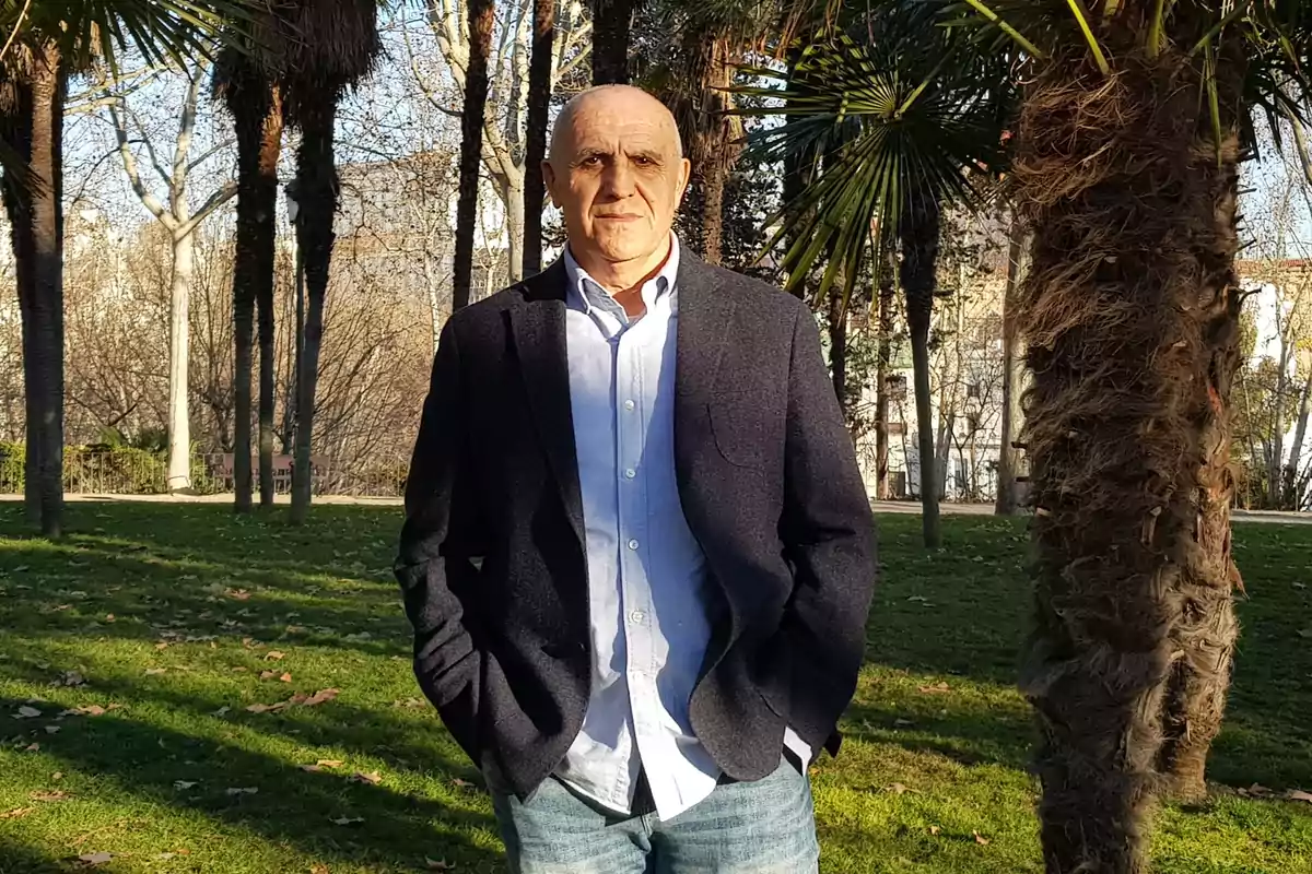 Hombre de mediana edad con chaqueta oscura y camisa clara posando en un parque con césped y palmeras en un día soleado Hombre de mediana edad con chaqueta oscura y camisa clara posando en un parque con césped y palmeras en un día soleado