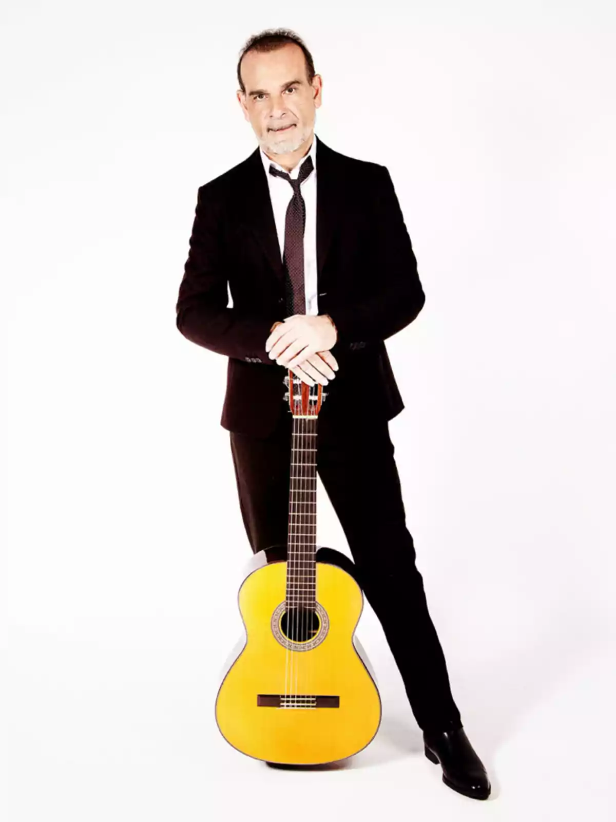 Hombre de traje oscuro y corbata posando de pie con una guitarra clásica apoyada en el suelo sobre fondo blanco Hombre de traje oscuro y corbata posando de pie con una guitarra clásica apoyada en el suelo sobre fondo blanco