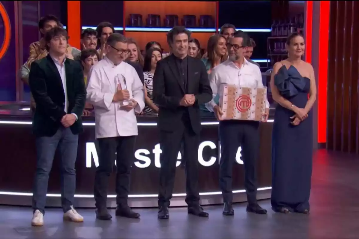 Grupo de personas -que son el jurado de MasterChef- en un set de televisión de un concurso de cocina, algunos sostienen un trofeo y una caja con el logo del programa, mientras el resto aplaude y sonríe en el fondo.