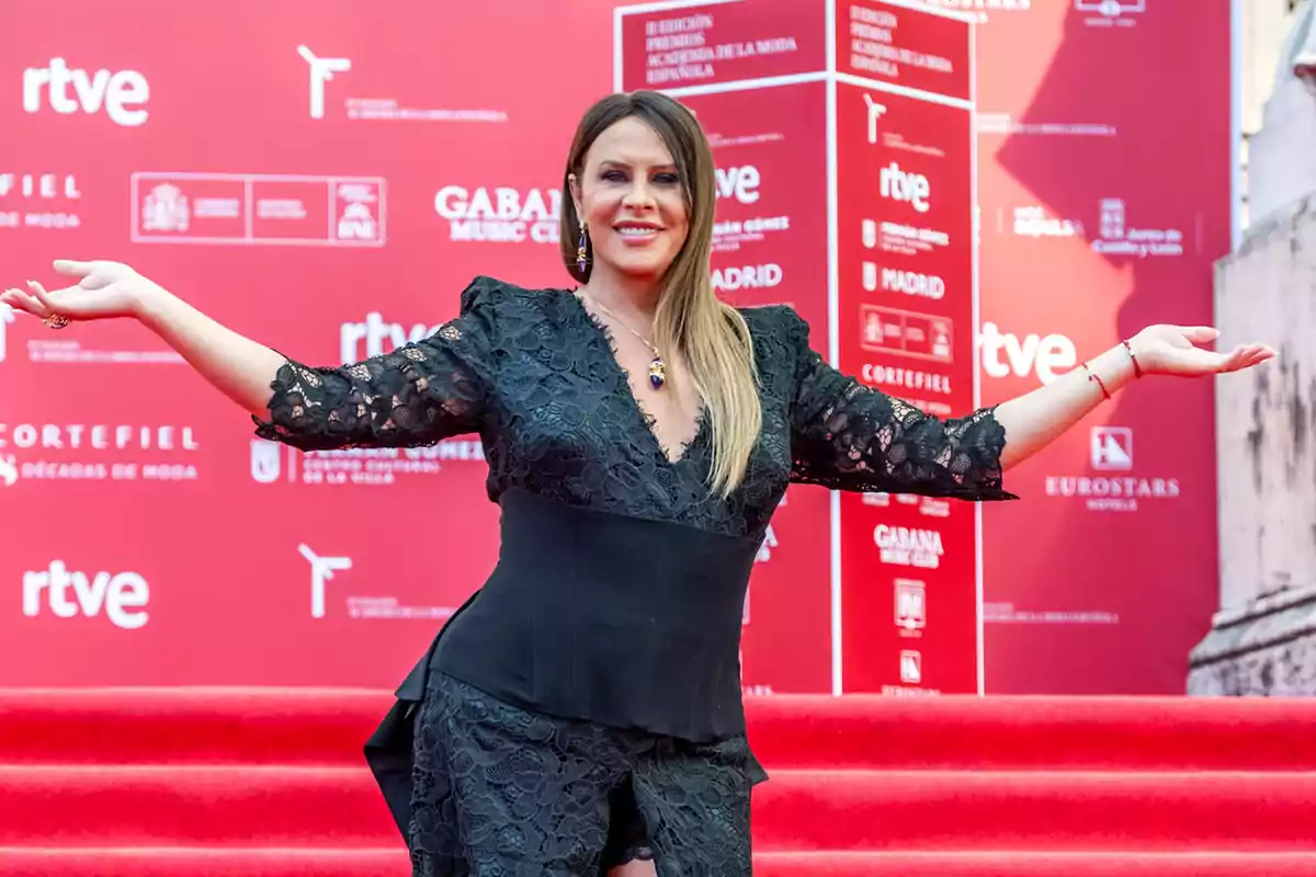 Mujer con vestido negro de encaje posando sonriente con los brazos abiertos sobre una alfombra roja en un evento