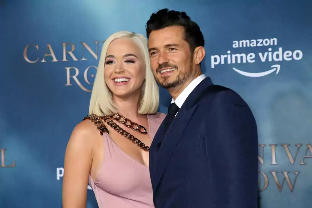 Una pareja sonríe posando en la alfombra azul de un evento con fondo de Amazon Prime Video y Carnival Row Una pareja sonríe posando en la alfombra azul de un evento con fondo de Amazon Prime Video y Carnival Row