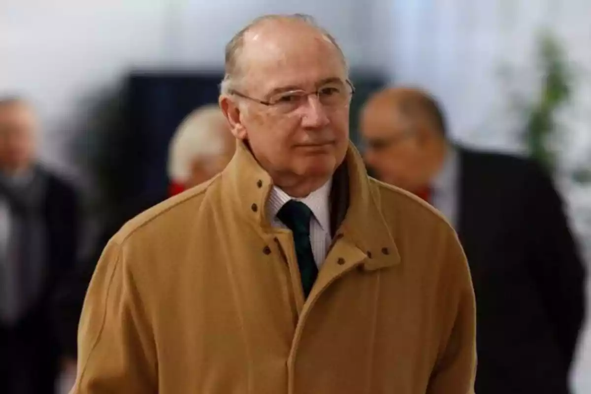 Hombre mayor con gafas y abrigo beige caminando en un entorno interior con personas desenfocadas al fondo Hombre mayor con gafas y abrigo beige caminando en un entorno interior con personas desenfocadas al fondo