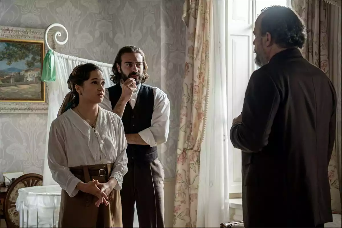 Catalina, Adriano y Alonso conversan en una habitación elegante con cortinas y papel tapiz, dos de ellas miran atentamente a un hombre de espaldas junto a una ventana iluminada. Captura de 'La Promesa' Catalina, Adriano y Alonso conversan en una habitación elegante con cortinas y papel tapiz, dos de ellas miran atentamente a un hombre de espaldas junto a una ventana iluminada. Captura de 'La Promesa'