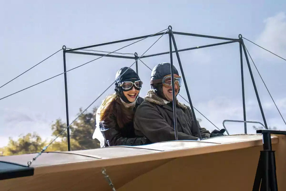 Enora y Manuel sonrientes con gafas y gorros de aviador disfrutan un vuelo en un avión antiguo bajo un cielo despejado. Captura de 'La Promesa' Enora y Manuel sonrientes con gafas y gorros de aviador disfrutan un vuelo en un avión antiguo bajo un cielo despejado. Captura de 'La Promesa'