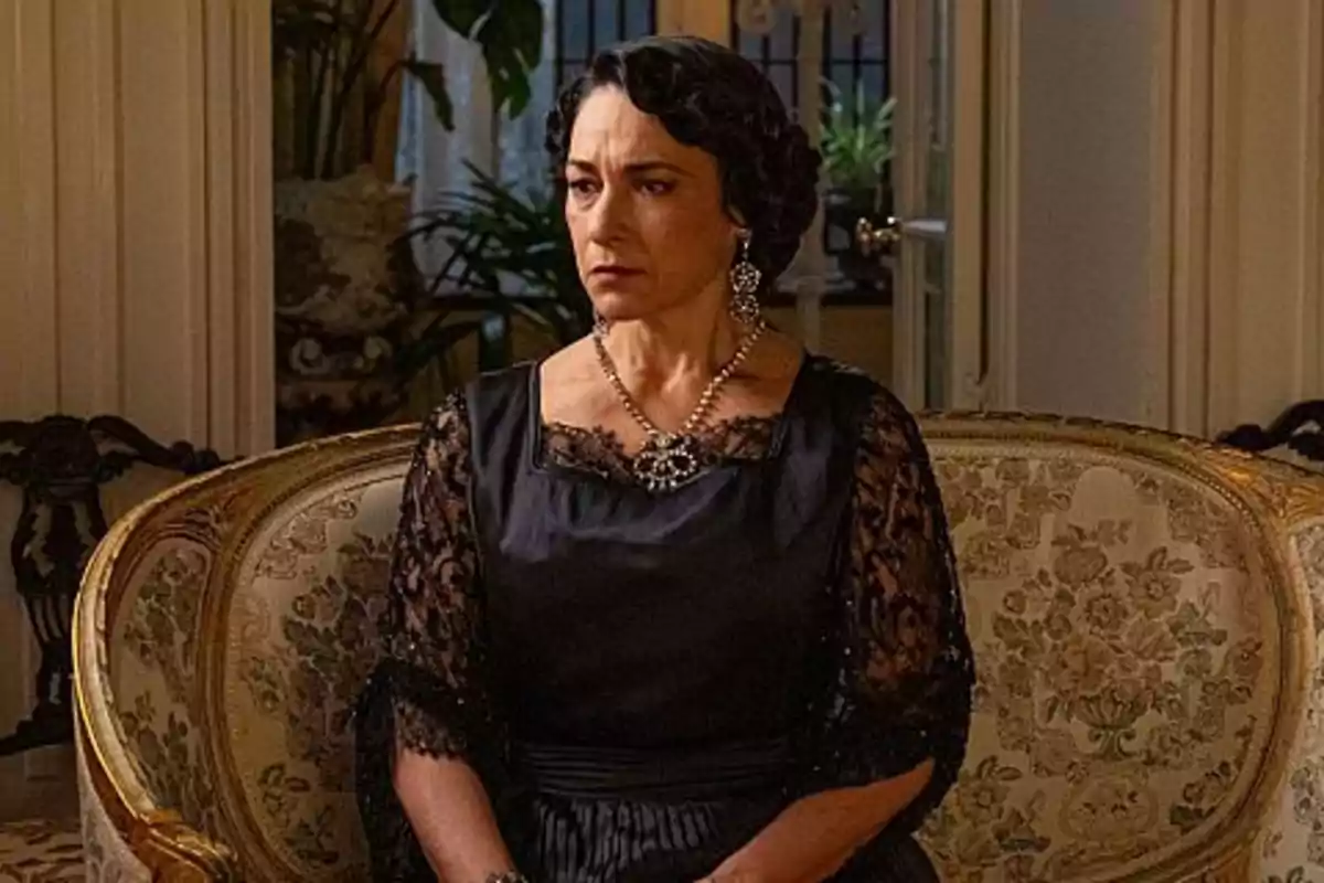 Leocadia de expresión seria sentada en un sofá antiguo con vestido negro elegante y joyas llamativas en una sala decorada con plantas y muebles clásicos. Captura de 'La Promesa' Leocadia de expresión seria sentada en un sofá antiguo con vestido negro elegante y joyas llamativas en una sala decorada con plantas y muebles clásicos. Captura de 'La Promesa'