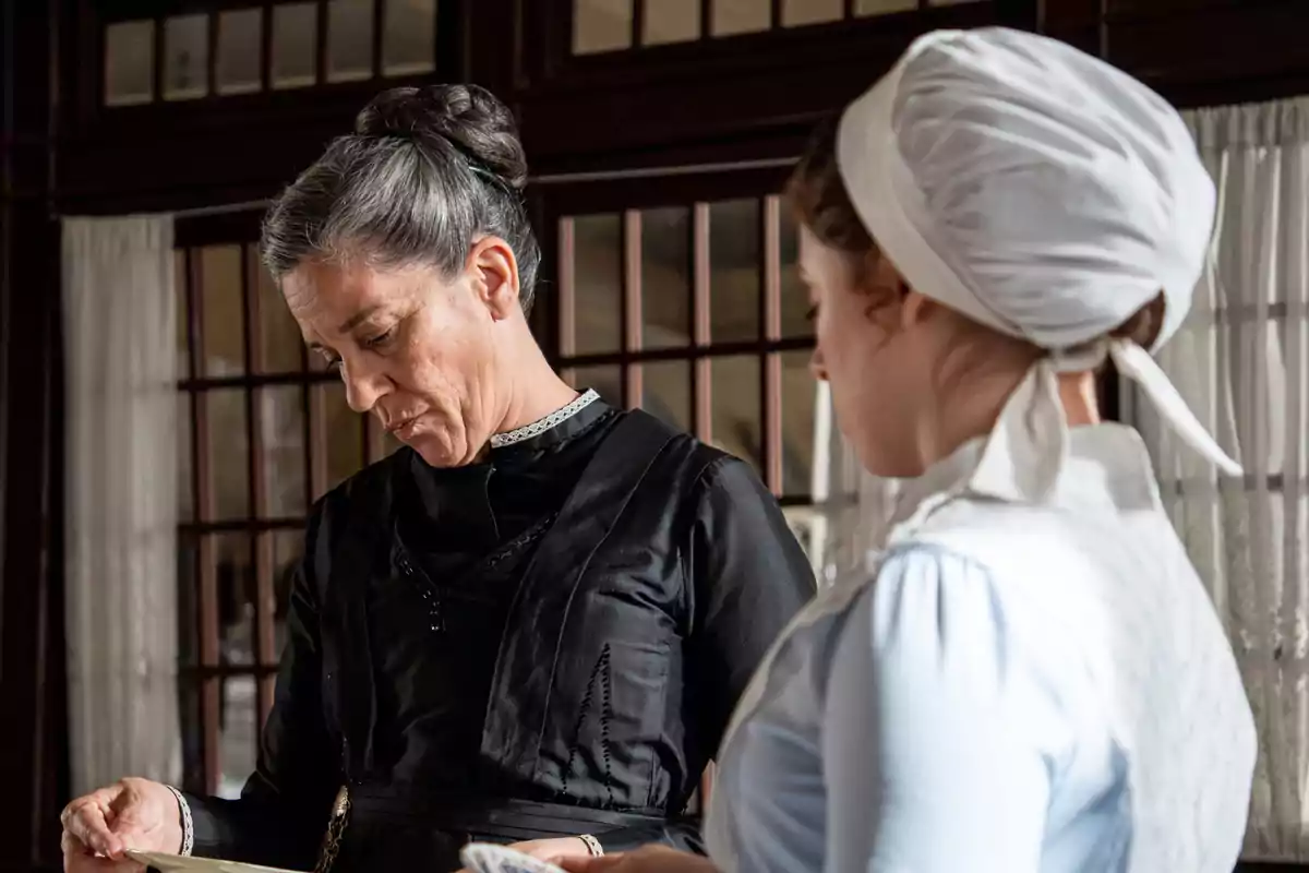 Marga y María Fernández con vestimenta de época están en una habitación con ventanas grandes y cortinas blancas, una de ellas lleva un vestido negro y la otra un gorro blanco. Captura de 'La Promesa' Marga y María Fernández con vestimenta de época están en una habitación con ventanas grandes y cortinas blancas, una de ellas lleva un vestido negro y la otra un gorro blanco. Captura de 'La Promesa'