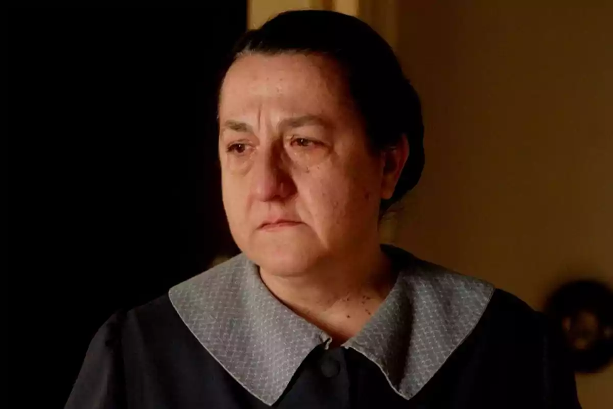 Simona con expresión seria y triste vestida con uniforme oscuro y cuello claro en un ambiente interior con luz cálida. Captura de 'La Promesa' Simona con expresión seria y triste vestida con uniforme oscuro y cuello claro en un ambiente interior con luz cálida. Captura de 'La Promesa'