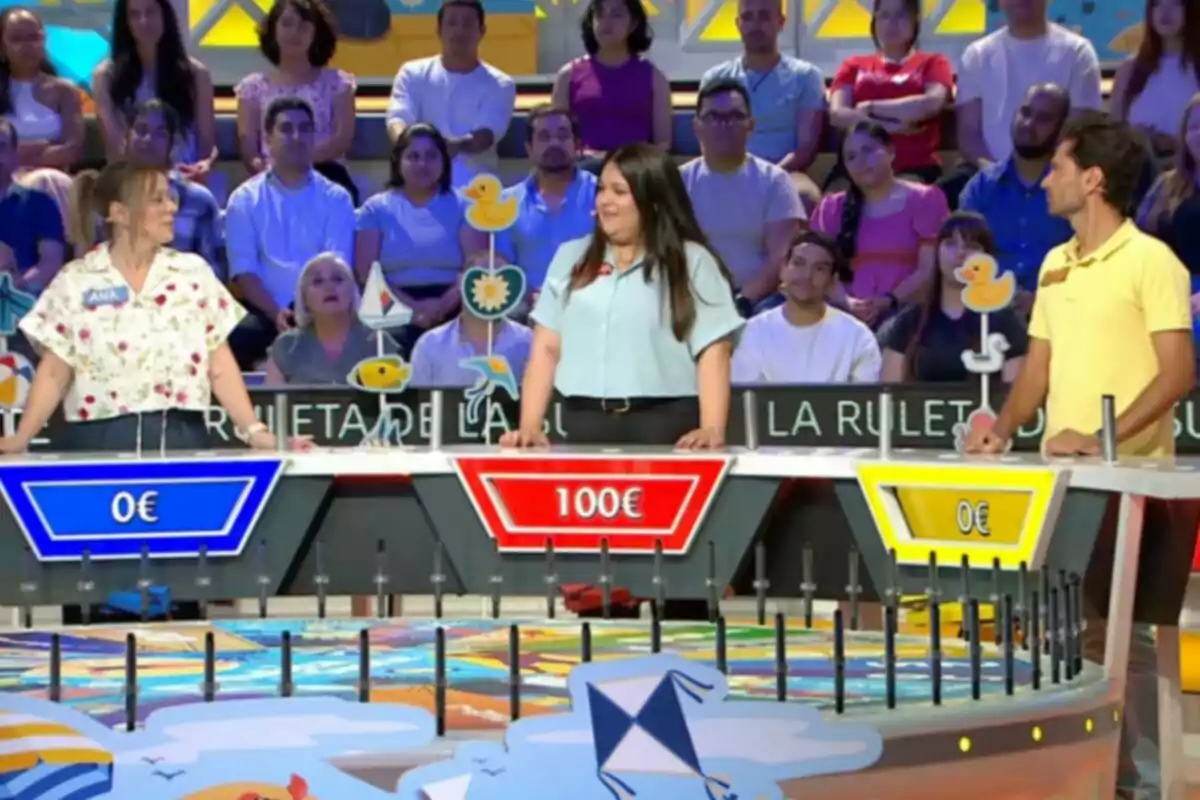Tres concursantes participan en un programa de juegos de televisión con un público de fondo y un panel de premios donde la concursante del centro lleva la delantera con 100 euros. Captura de 'La Ruleta de la Suerte' Tres concursantes participan en un programa de juegos de televisión con un público de fondo y un panel de premios donde la concursante del centro lleva la delantera con 100 euros. Captura de 'La Ruleta de la Suerte'