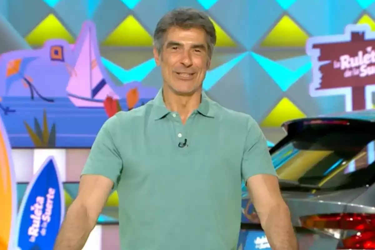 Jorge Fernández con camiseta verde claro sonríe en un set colorido de televisión con decoraciones y un coche al fondo. Captura de 'La Ruleta de la Suerte' Jorge Fernández con camiseta verde claro sonríe en un set colorido de televisión con decoraciones y un coche al fondo. Captura de 'La Ruleta de la Suerte'