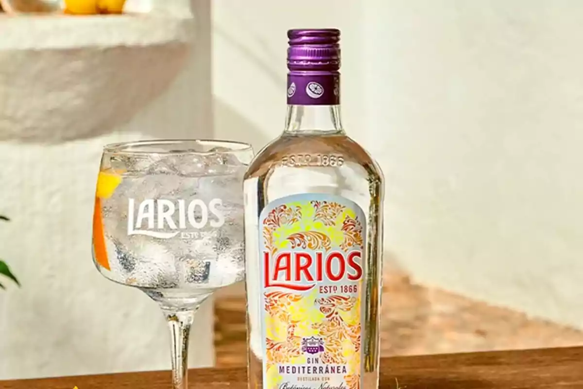 Botella de ginebra Larios junto a una copa con hielo y rodajas de cítricos sobre una mesa de madera en un ambiente luminoso Botella de ginebra Larios junto a una copa con hielo y rodajas de cítricos sobre una mesa de madera en un ambiente luminoso
