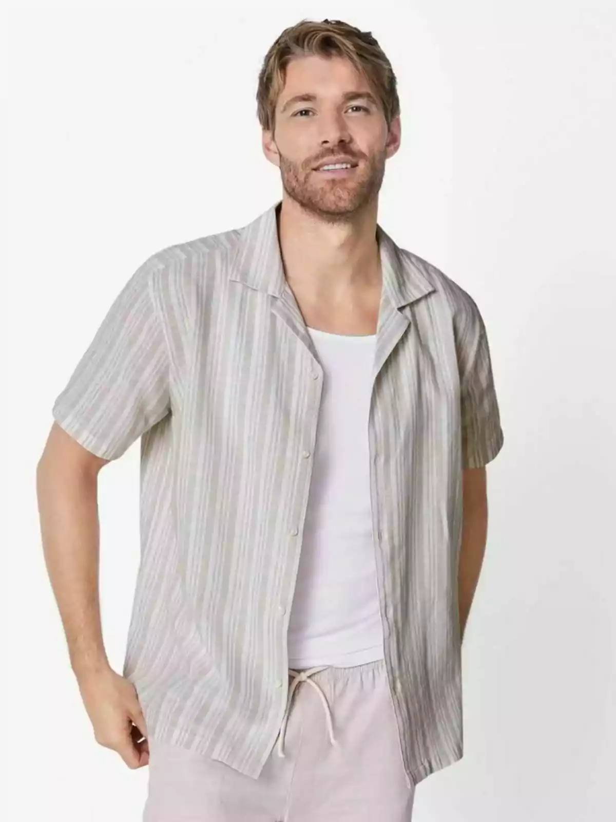 Hombre joven con barba y cabello rubio viste una camisa de manga corta a rayas sobre una camiseta blanca y pantalones claros posando frente a un fondo blanco Hombre joven con barba y cabello rubio viste una camisa de manga corta a rayas sobre una camiseta blanca y pantalones claros posando frente a un fondo blanco