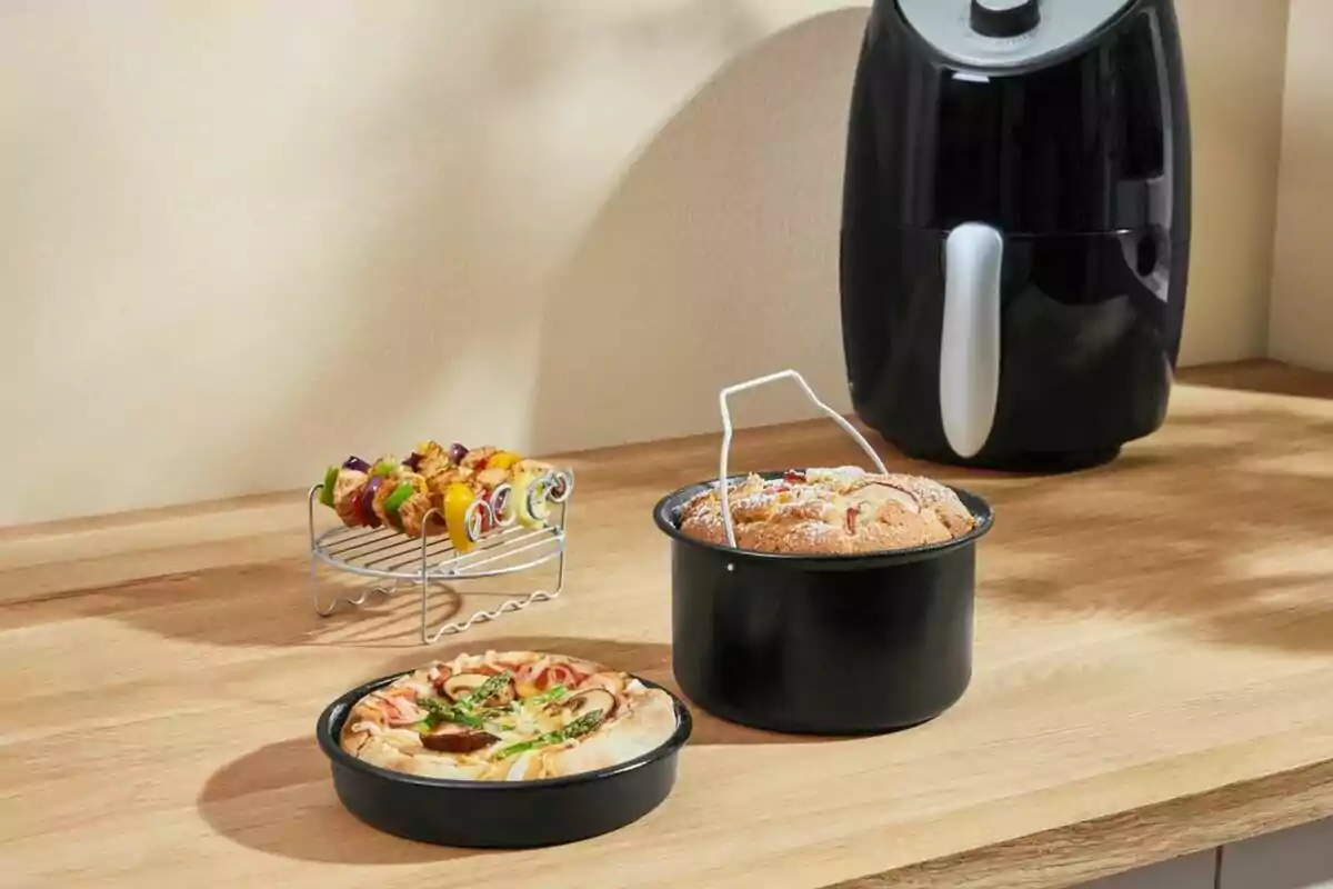Freidora de aire negra sobre una encimera de madera junto a un pastel, una pizza y brochetas de verduras y carne Freidora de aire negra sobre una encimera de madera junto a un pastel, una pizza y brochetas de verduras y carne