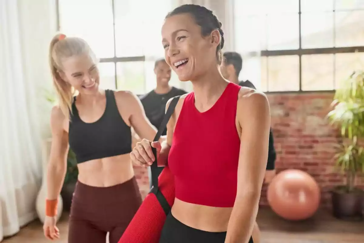 Mujeres jóvenes sonrientes con ropa deportiva en un estudio de yoga iluminado por la luz natural, una de ellas lleva un tapete rojo. Mujeres jóvenes sonrientes con ropa deportiva en un estudio de yoga iluminado por la luz natural, una de ellas lleva un tapete rojo.