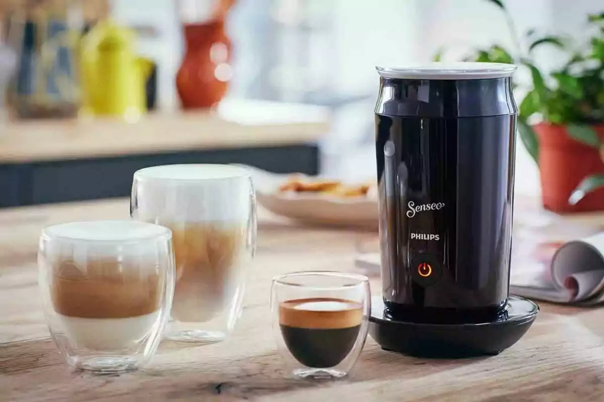 Espumador de leche eléctrico negro junto a tres vasos de café con diferentes niveles de espuma sobre una mesa de madera en una cocina iluminada Espumador de leche eléctrico negro junto a tres vasos de café con diferentes niveles de espuma sobre una mesa de madera en una cocina iluminada
