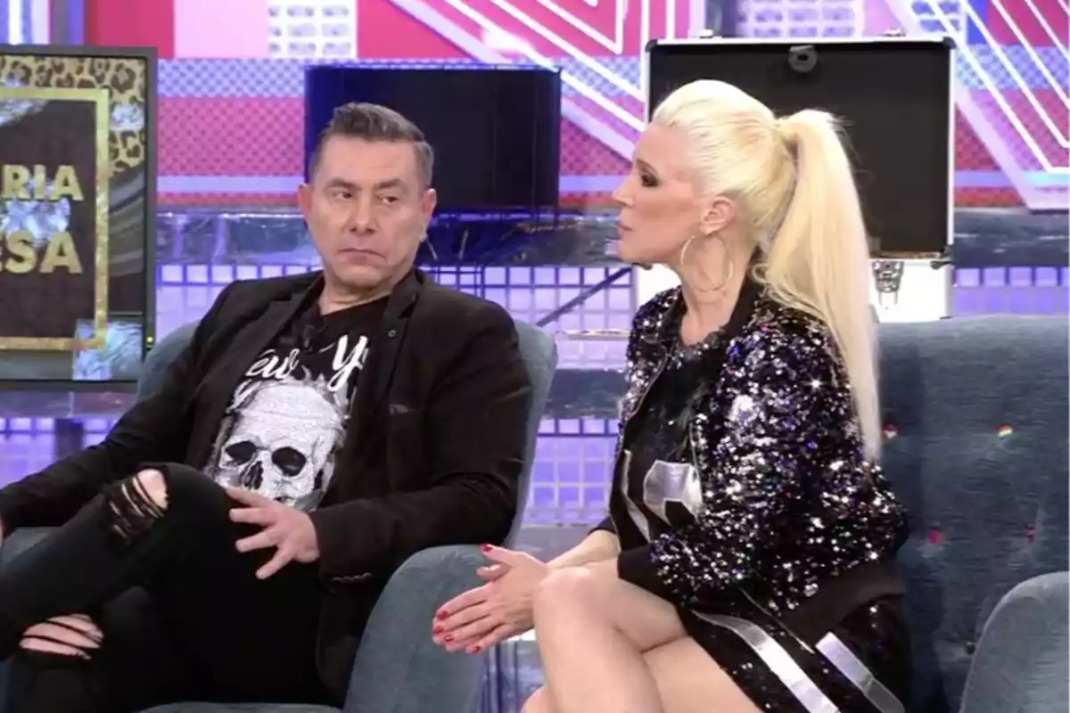 Dos personas sentadas en un set de televisión conversando, una mujer rubia con coleta y chaqueta de lentejuelas y un hombre con camiseta de calavera y pantalón roto Dos personas sentadas en un set de televisión conversando, una mujer rubia con coleta y chaqueta de lentejuelas y un hombre con camiseta de calavera y pantalón roto