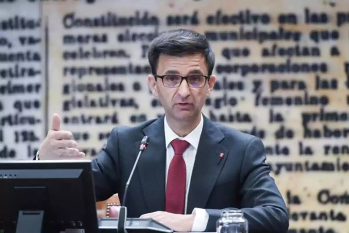 Hombre con gafas y traje oscuro hablando en una conferencia frente a un fondo con texto borroso Hombre con gafas y traje oscuro hablando en una conferencia frente a un fondo con texto borroso