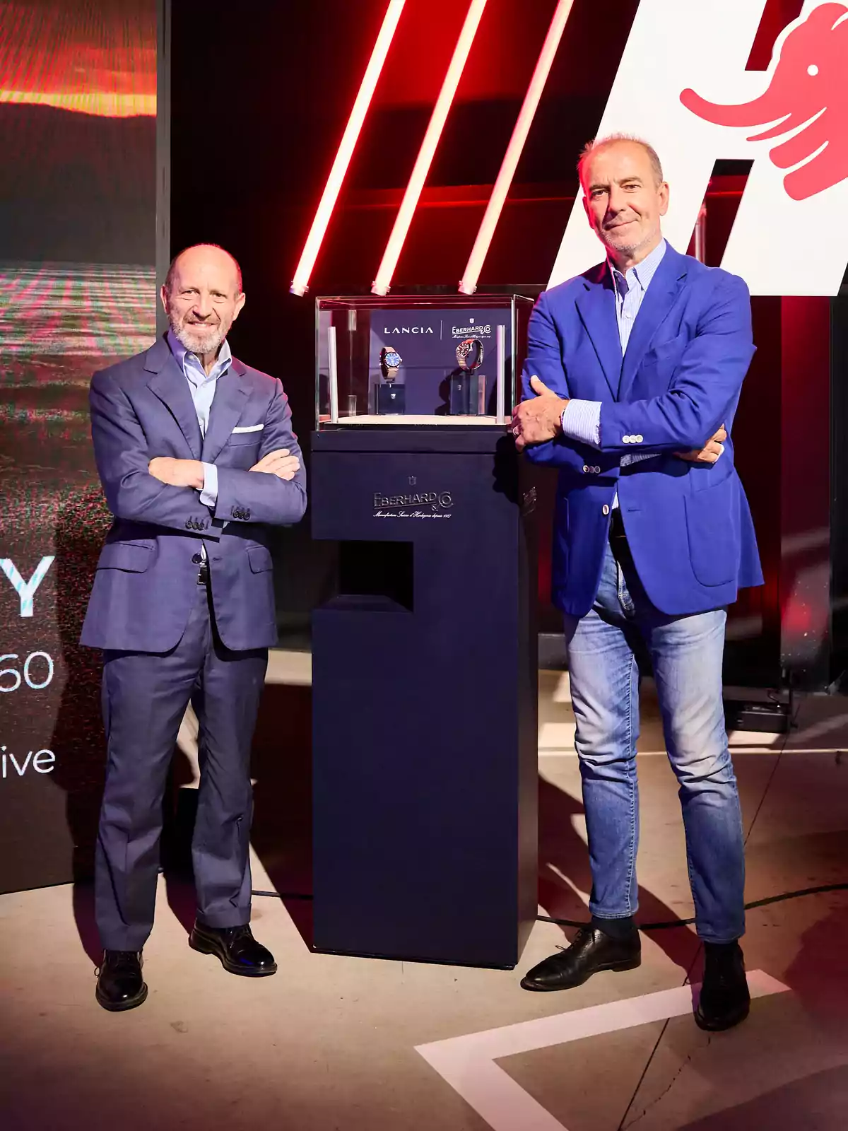 Dos hombres posan sonrientes junto a una vitrina que exhibe relojes en un evento elegante, ambos visten saco azul y camisas claras Dos hombres posan sonrientes junto a una vitrina que exhibe relojes en un evento elegante, ambos visten saco azul y camisas claras