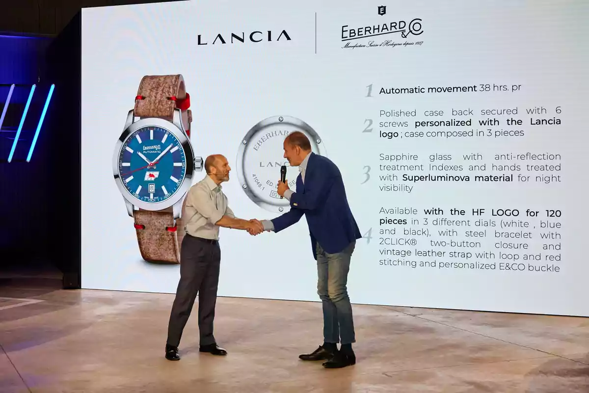 Dos hombres se dan la mano frente a una pantalla grande que muestra un reloj de pulsera y detalles sobre sus características, junto a los logotipos de Lancia y Eberhard & Co.