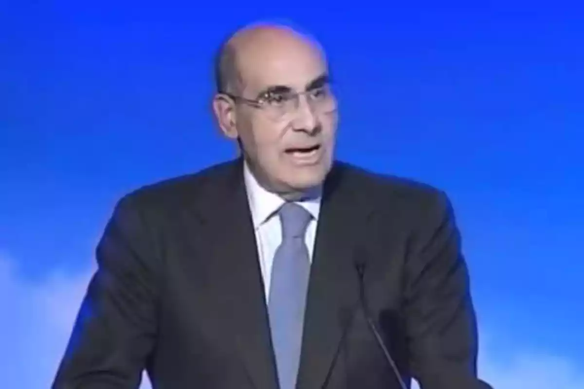 Hombre calvo con gafas y traje oscuro hablando frente a un micrófono con fondo azul Hombre calvo con gafas y traje oscuro hablando frente a un micrófono con fondo azul