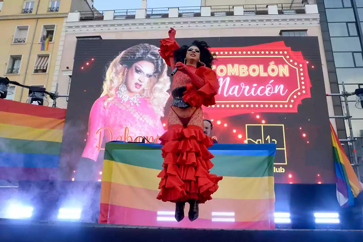 Drag queen vestida con un llamativo traje rojo actúa en un escenario decorado con banderas del orgullo LGBT y una gran pantalla de fondo en un evento al aire libre Drag queen vestida con un llamativo traje rojo actúa en un escenario decorado con banderas del orgullo LGBT y una gran pantalla de fondo en un evento al aire libre