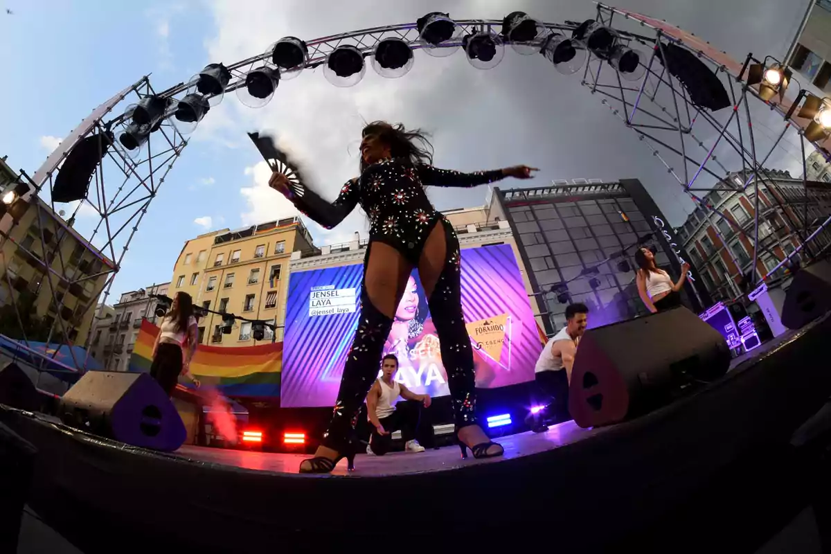 Una persona con vestuario llamativo y brillante baila en un escenario al aire libre durante un evento con luces de colores y una pantalla gigante de fondo, acompañada de otros bailarines y con una bandera arcoíris visible entre los edificios.