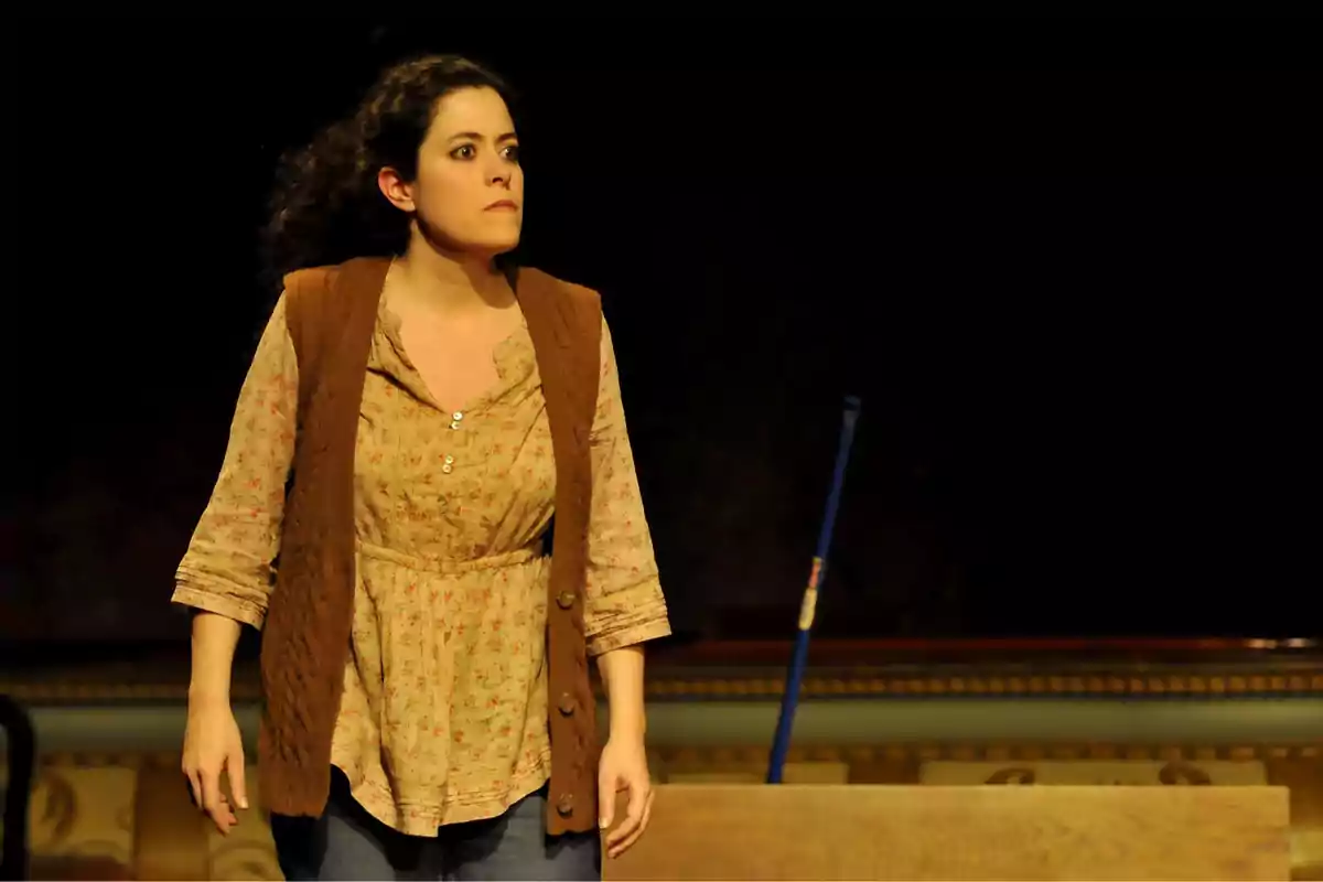 Mujer de cabello rizado y expresión seria vestida con blusa floreada y chaleco marrón en un escenario teatral con fondo oscuro y un palo azul detrás de ella