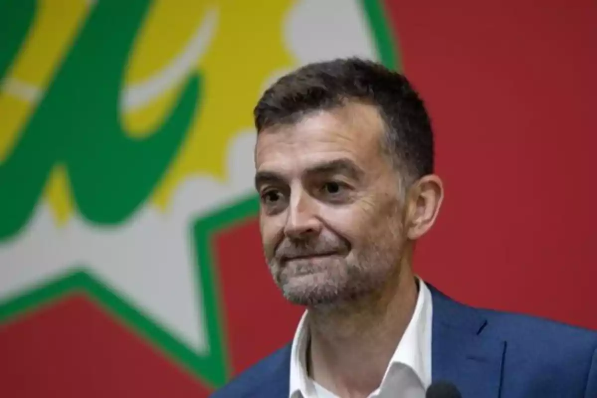 Hombre de cabello corto y barba con saco azul y camisa blanca sonriendo frente a un fondo colorido con formas verdes, amarillas y rojas Hombre de cabello corto y barba con saco azul y camisa blanca sonriendo frente a un fondo colorido con formas verdes, amarillas y rojas