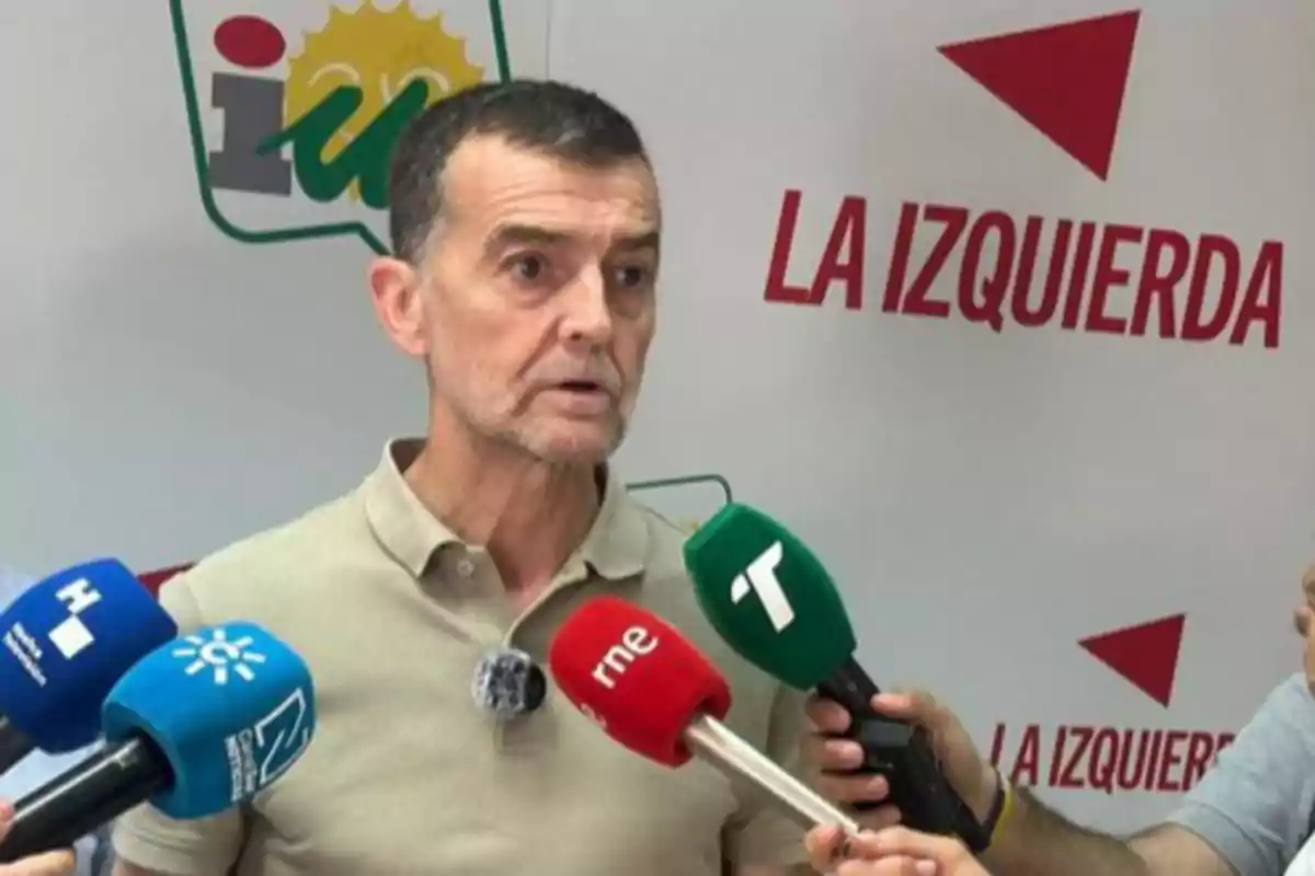 Un hombre de cabello corto y barba recortada responde preguntas de varios medios de comunicación frente a un fondo con el logo de Izquierda Unida y el texto La Izquierda Un hombre de cabello corto y barba recortada responde preguntas de varios medios de comunicación frente a un fondo con el logo de Izquierda Unida y el texto La Izquierda