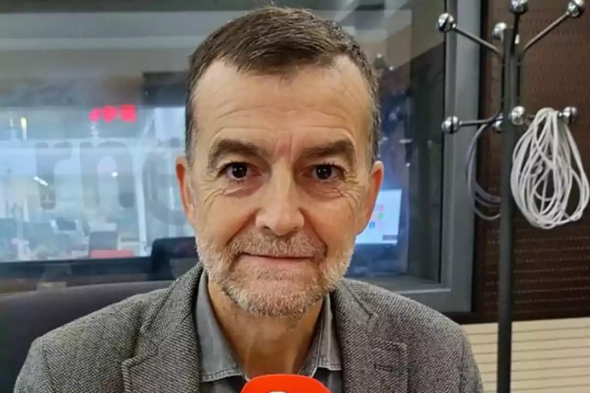 Hombre de cabello corto y barba canosa con chaqueta gris sosteniendo un micrófono rojo en un estudio de radio Hombre de cabello corto y barba canosa con chaqueta gris sosteniendo un micrófono rojo en un estudio de radio