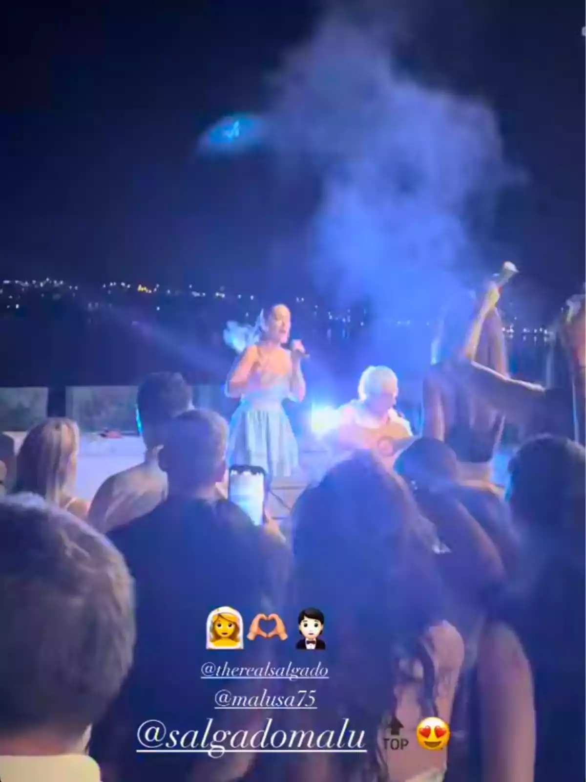 Mujer cantando en un escenario al aire libre durante la noche frente a un público, con luces azules y humo iluminando el ambiente Mujer cantando en un escenario al aire libre durante la noche frente a un público, con luces azules y humo iluminando el ambiente