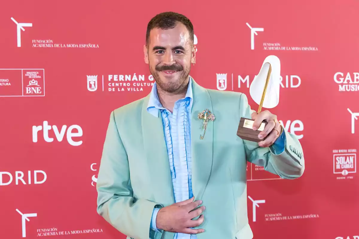 Hombre con bigote y chaqueta verde claro sonríe mientras sostiene un trofeo frente a un fondo rojo con logotipos de patrocinadores. Hombre con bigote y chaqueta verde claro sonríe mientras sostiene un trofeo frente a un fondo rojo con logotipos de patrocinadores.