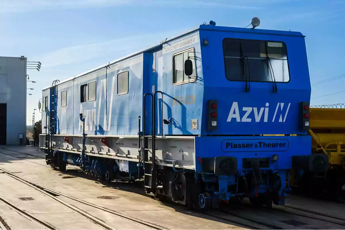 Vagón ferroviario azul de mantenimiento con la marca Azvi estacionado sobre las vías junto a una nave industrial Vagón ferroviario azul de mantenimiento con la marca Azvi estacionado sobre las vías junto a una nave industrial