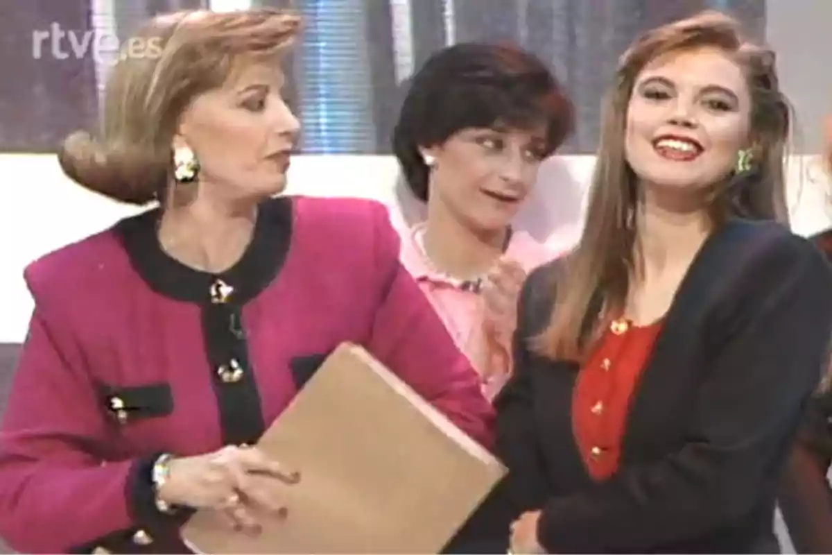 Tres mujeres vestidas con ropa elegante de los años 90 en un set de televisión, una de ellas sostiene una carpeta y otra sonríe hacia la cámara Tres mujeres vestidas con ropa elegante de los años 90 en un set de televisión, una de ellas sostiene una carpeta y otra sonríe hacia la cámara