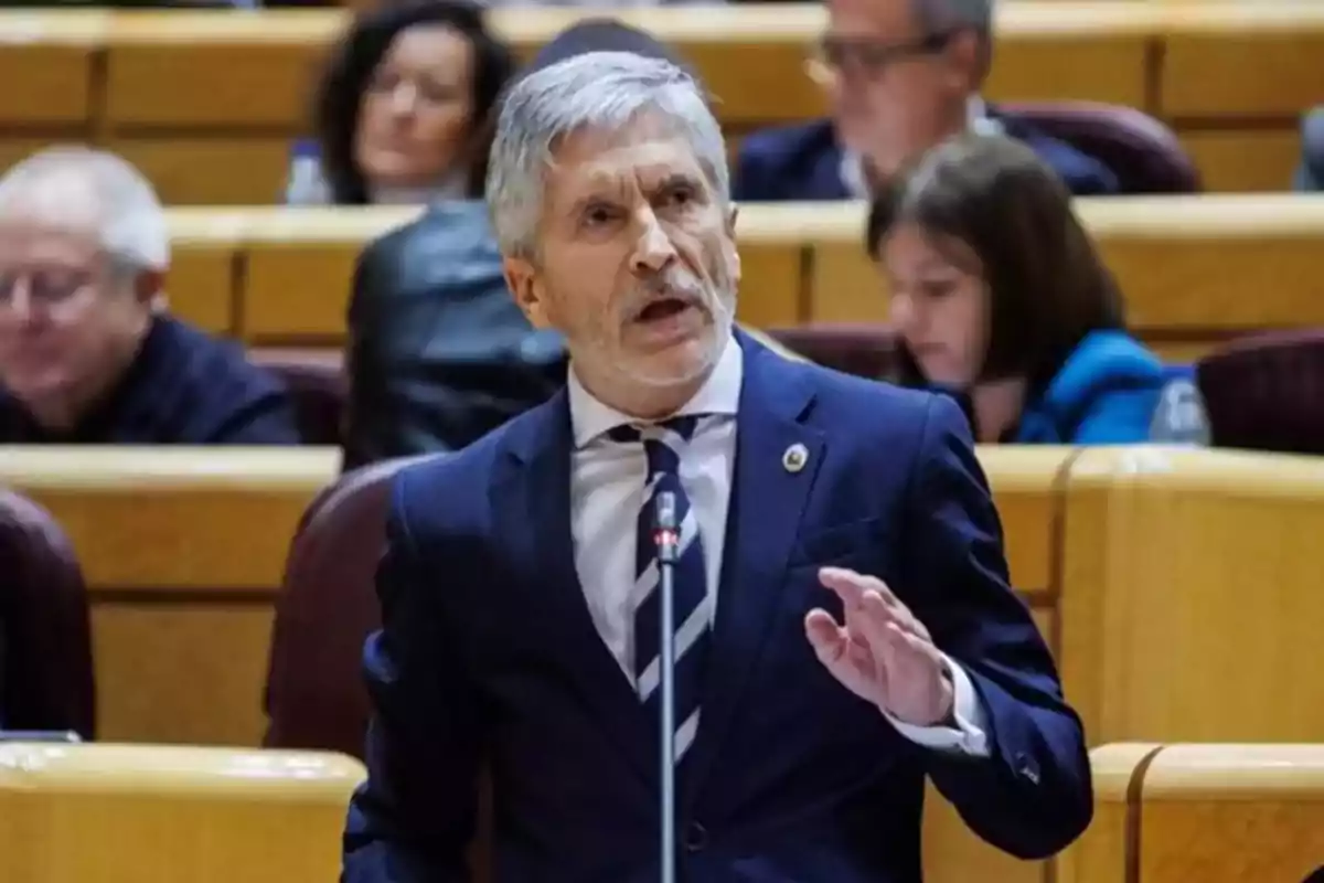 Hombre de cabello canoso y traje azul hablando en un micrófono en un recinto parlamentario con personas sentadas al fondo Hombre de cabello canoso y traje azul hablando en un micrófono en un recinto parlamentario con personas sentadas al fondo