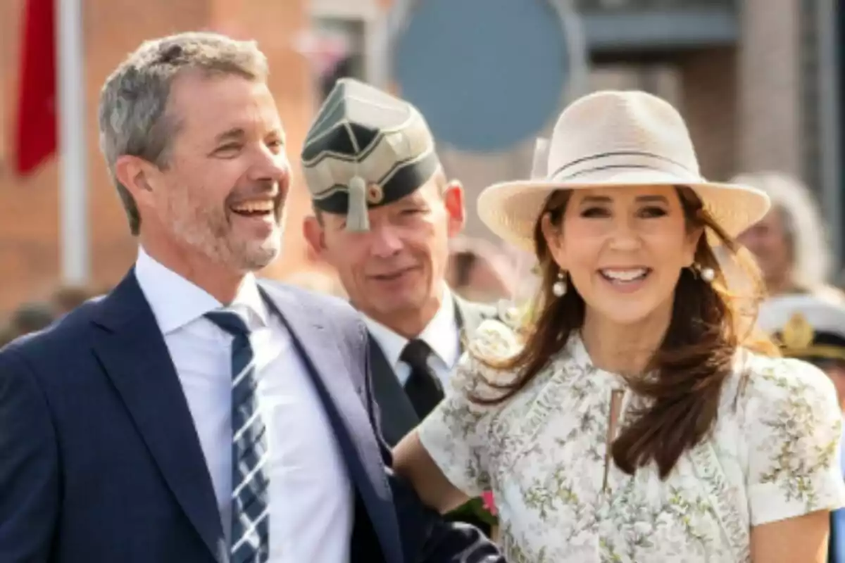 Pareja sonriente vestida de manera elegante -formada por Mary de Dinamarca y Federico de Dinamarca- en un evento al aire libre acompañada por un hombre con uniforme militar. Pareja sonriente vestida de manera elegante -formada por Mary de Dinamarca y Federico de Dinamarca- en un evento al aire libre acompañada por un hombre con uniforme militar.
