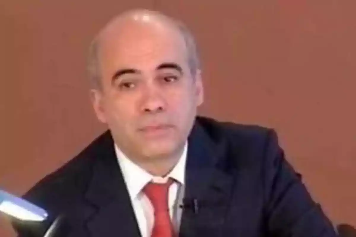 Hombre calvo con traje oscuro y corbata roja sentado frente a un fondo marrón